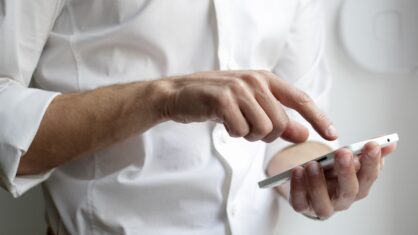 Weißes Smartphone in einer Hand, die vor einem Menschen in weißem Businesshemd gehalten wird.